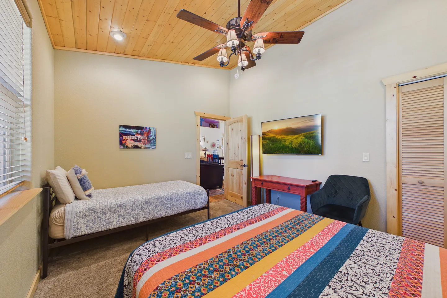 Bedroom with wood-plank ceiling, ceiling fan, twin bed, colorful larger bed, and wall-mounted TV.