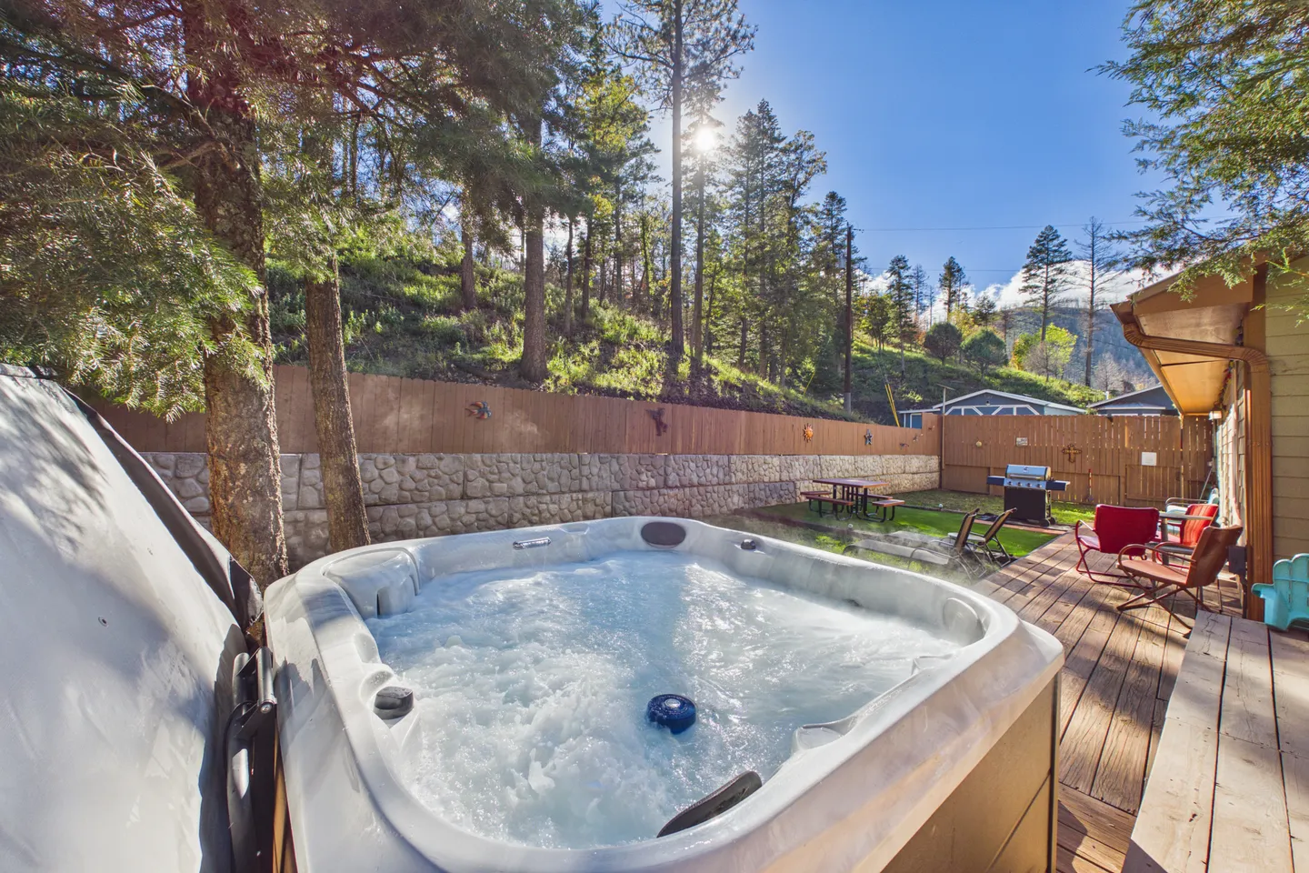 A bubbling outdoor hot tub on a wooden deck in a backyard surrounded by tall pine trees and a wooden fence.