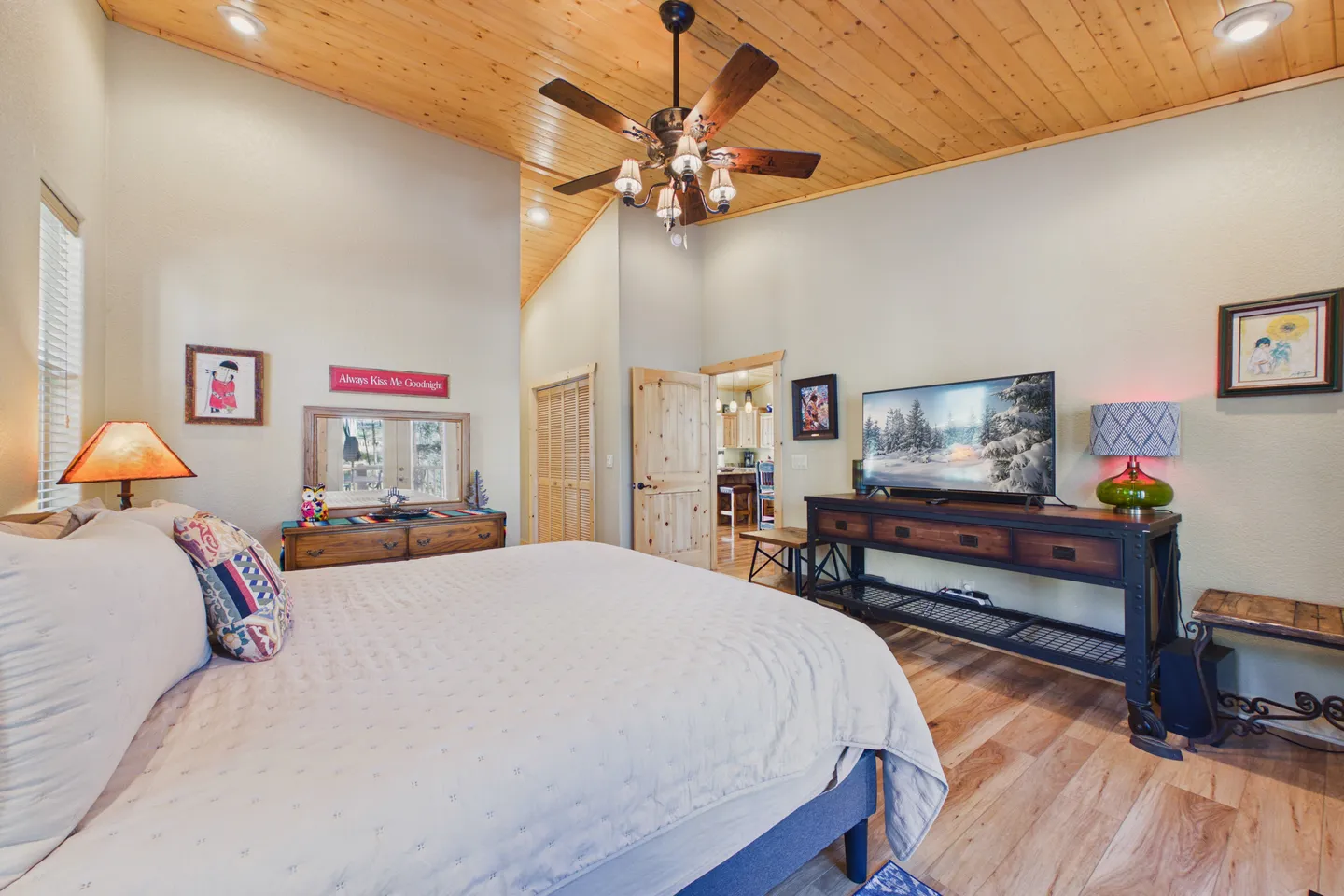 Bedroom with a wood-paneled angled ceiling, ceiling fan, made bed, and television on a console table.