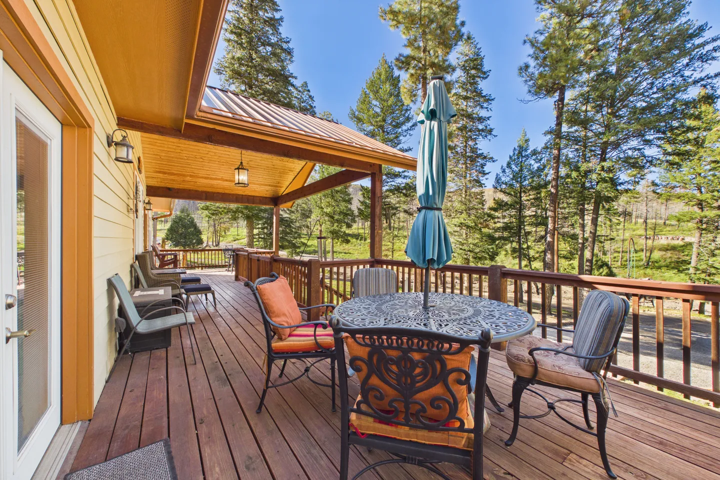 Spacious wooden outdoor deck featuring a patio dining table with chairs, an umbrella, and lounge seating beneath a covered section overlooking pine trees.