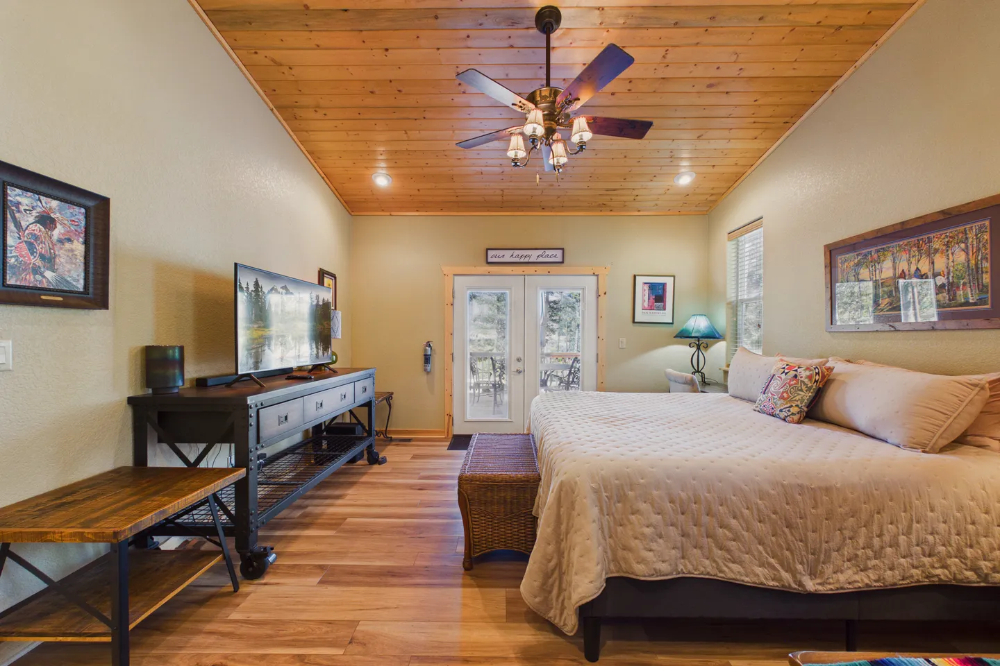 Bedroom with a wood-plank ceiling, ceiling fan, large bed, media console with TV, and glass doors leading outside.