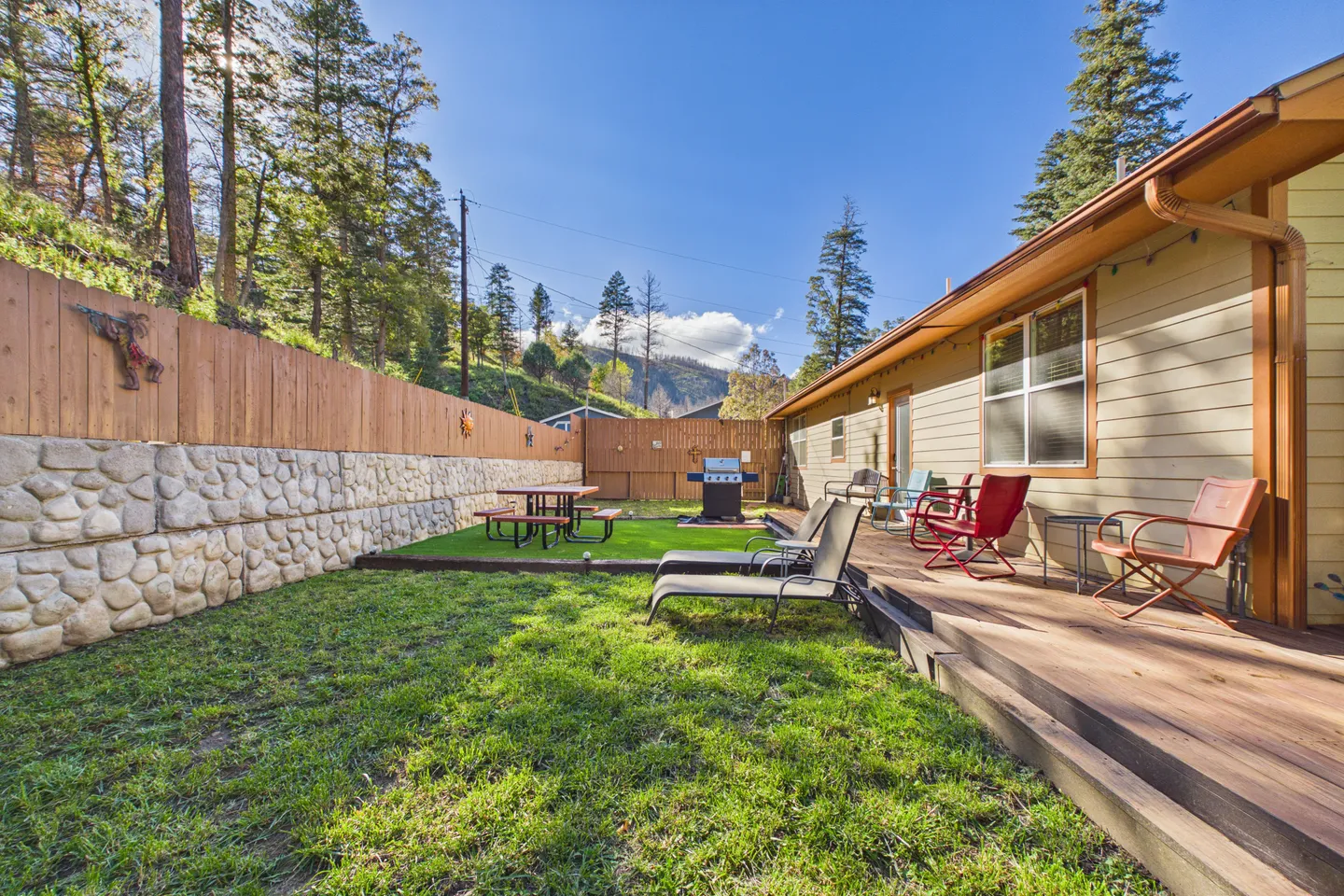 Fenced backyard featuring a wooden deck with outdoor chairs, a grass lawn, a stone retaining wall, a picnic table, and a grill.