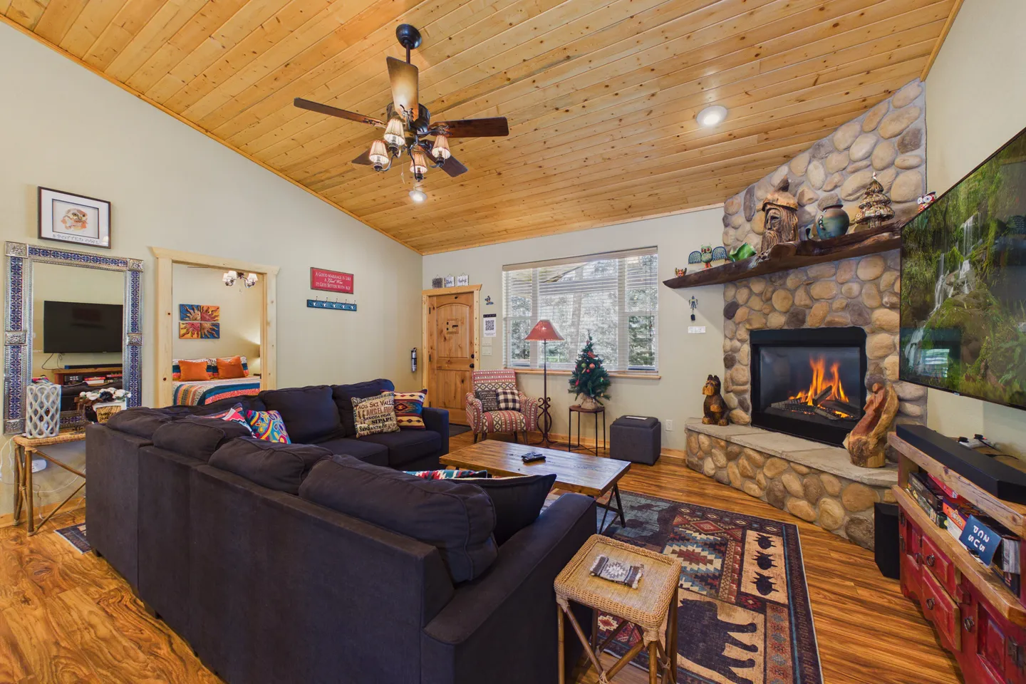 Living room featuring a dark sectional sofa, wood-plank vaulted ceiling with a fan, corner stone fireplace with an active fire, and a mounted TV.