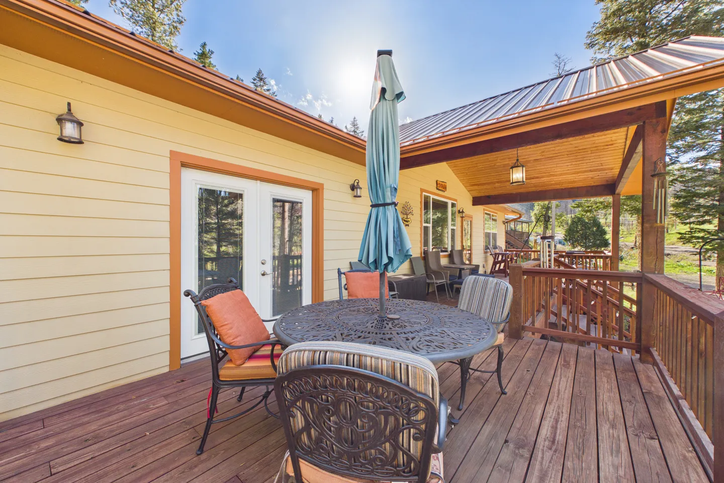 Outdoor wooden deck featuring a dining table with an umbrella, chairs with cushions, and railings next to a yellow house with a covered porch area.