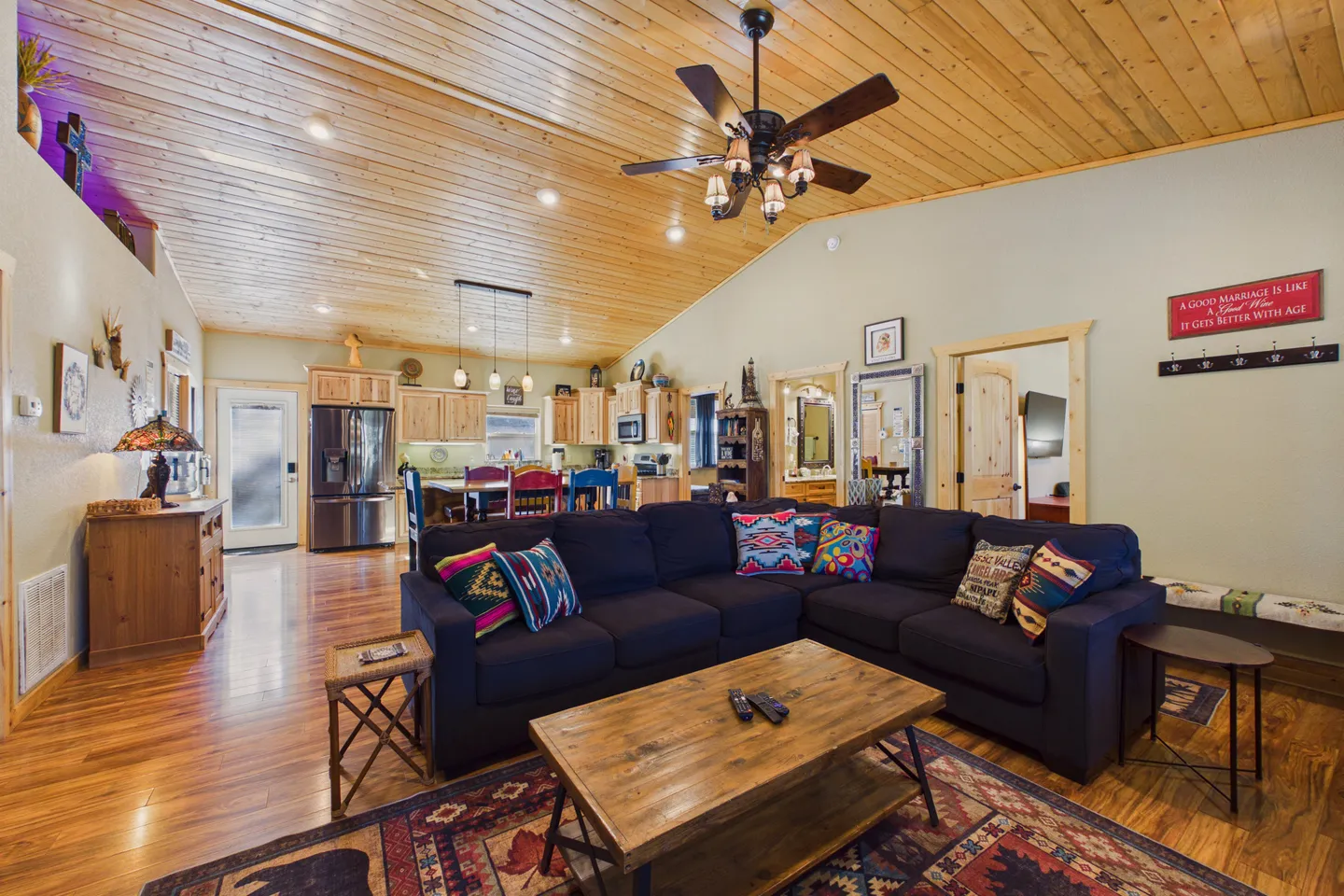 Open-concept living room with a dark sectional sofa, wood-paneled vaulted ceiling, and view into the kitchen.