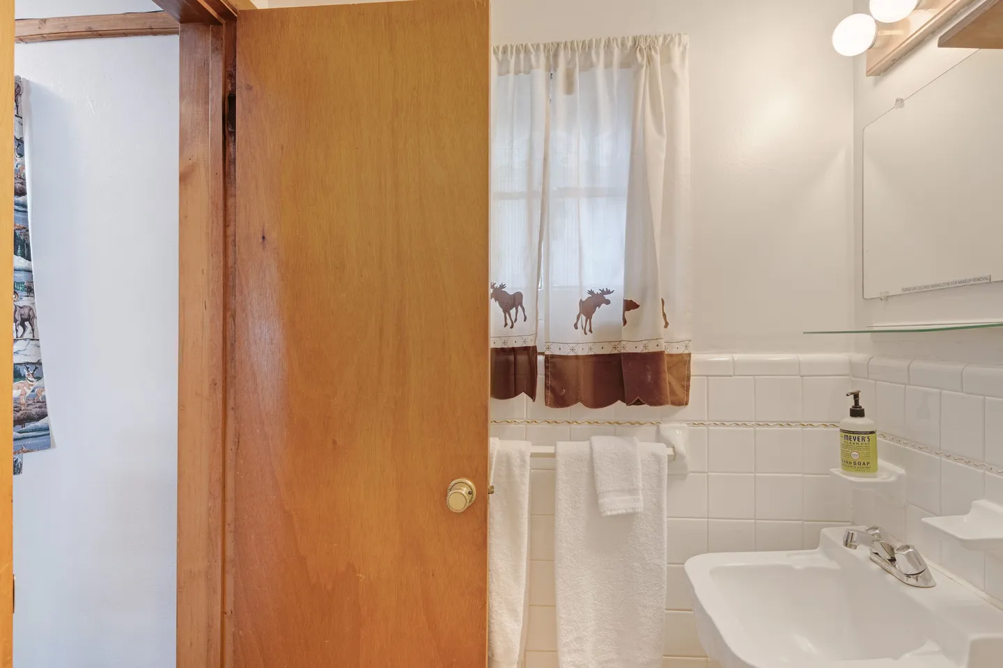 Bathroom with white tiled walls, a sink, a partially open wooden door, and a moose-themed window curtain.