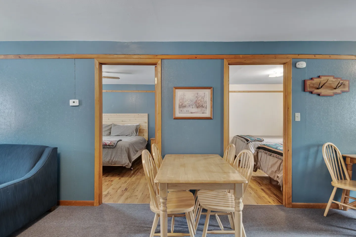 Dining area with a wooden table and chairs against a blue wall with doorways opening to two bedrooms.