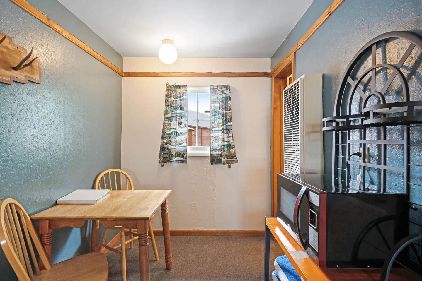 Dining nook with a wooden table, two wooden chairs, a window with curtains, and a microwave on a nearby stand.