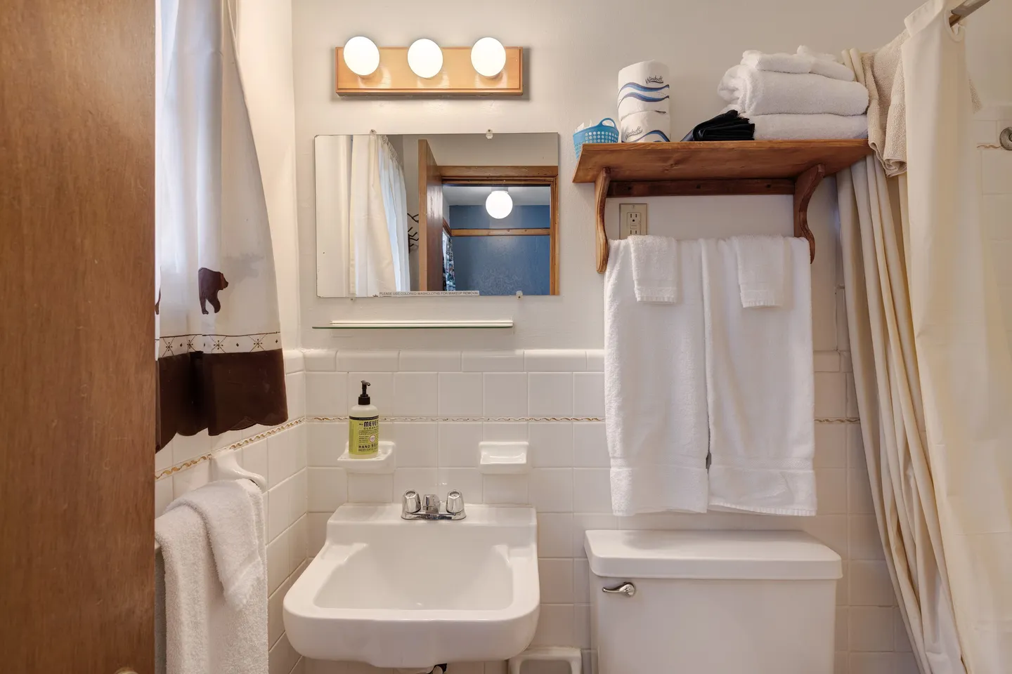 Bathroom with white wall-mounted sink, toilet, mirror with vanity lights, and a wooden shelf holding folded towels.