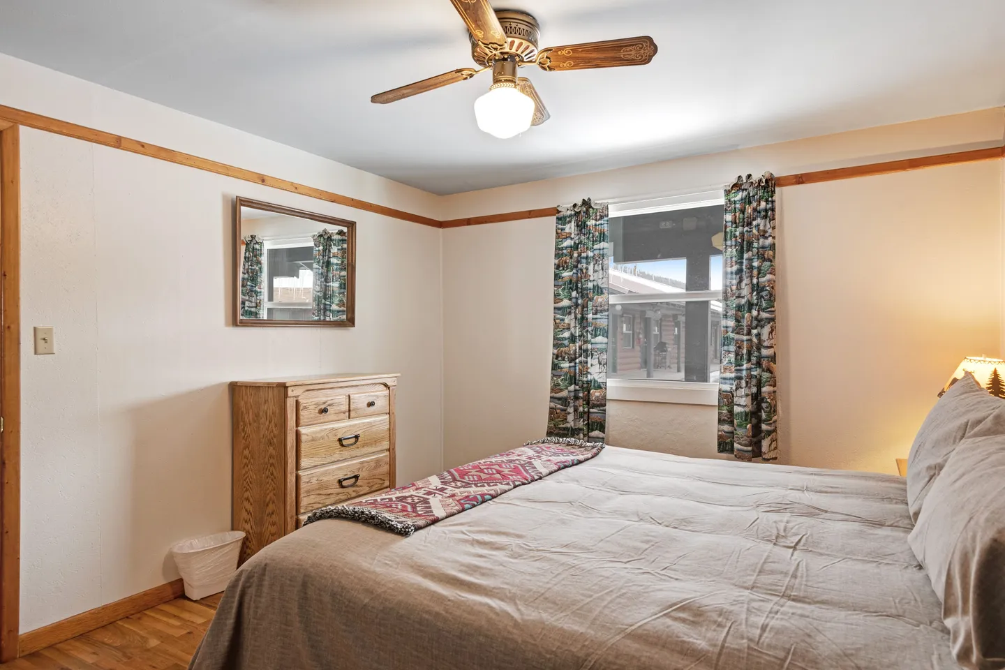 Bedroom featuring a made bed, wood dresser, wall mirror, and a window with patterned curtains below a ceiling fan.