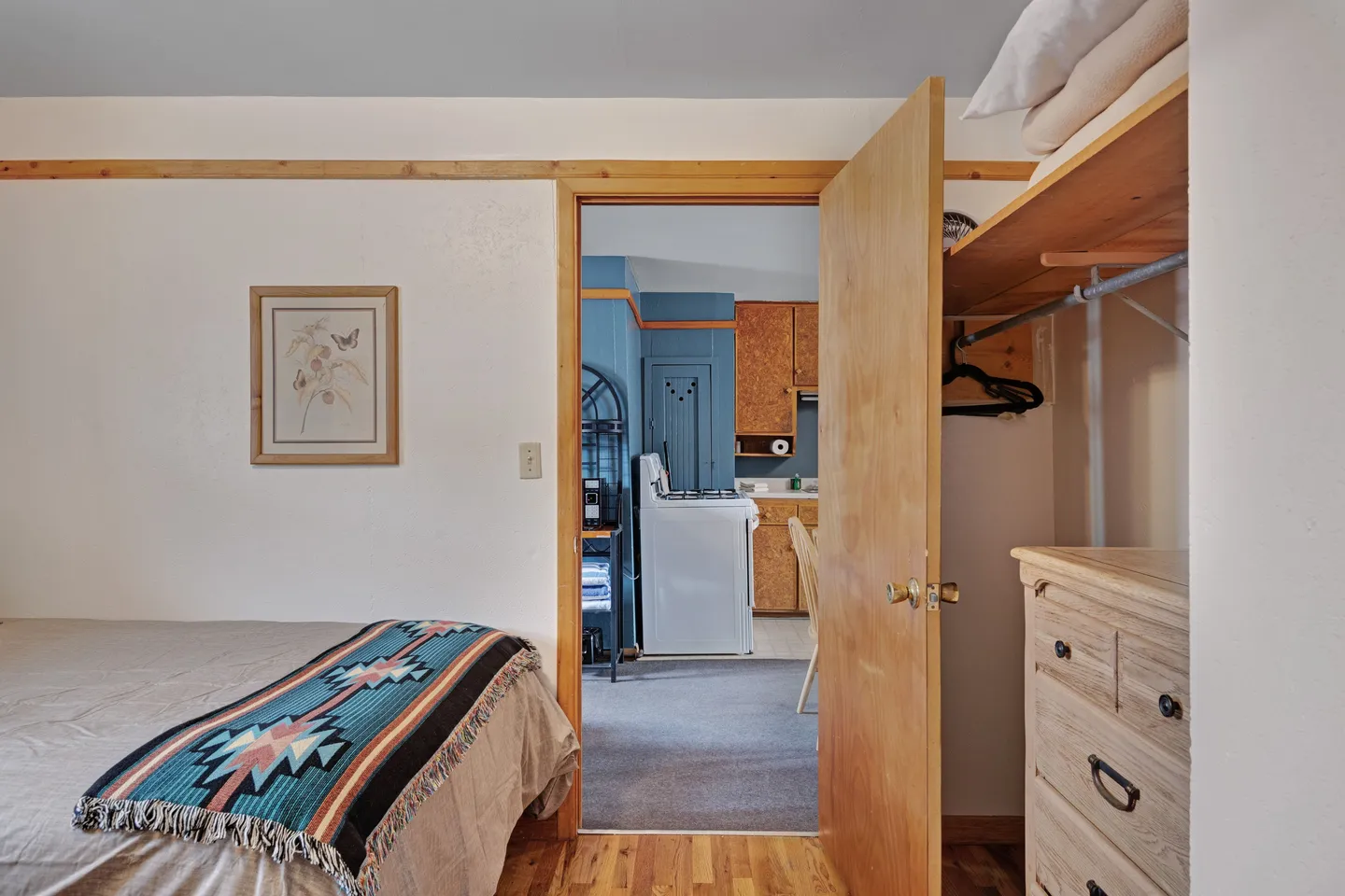 Bedroom with wooden floors and an open closet, looking through an open doorway into an adjacent kitchen.
