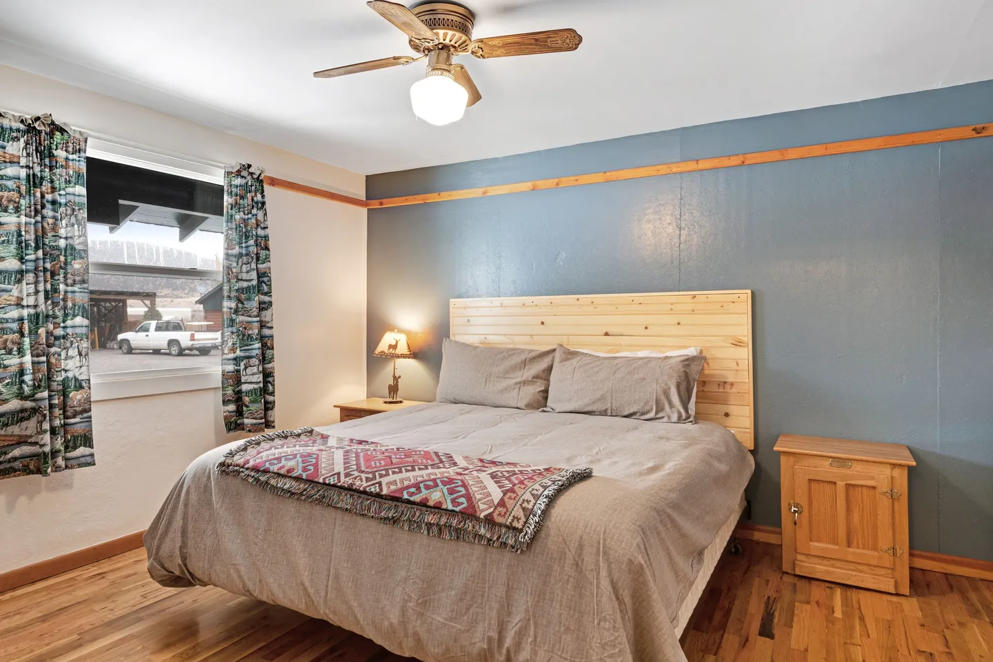 Bedroom featuring a made bed with a light wood headboard against a blue accent wall, a ceiling fan, and hardwood floors.