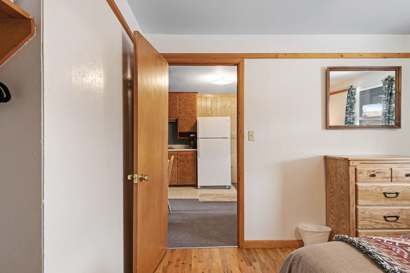 Open doorway from a bedroom with hardwood-look floors looking into a kitchen with wood cabinets and a white refrigerator.