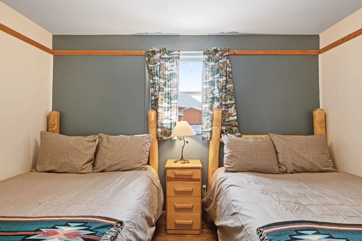 Bedroom with two beds, log headboards, a bedside table with a lamp, and a centered window with patterned curtains.