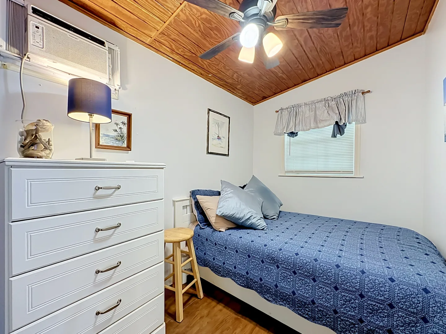 Bedroom with a sloped wood-plank ceiling, ceiling fan, single bed with blue bedspread, white dresser, and window air conditioner.