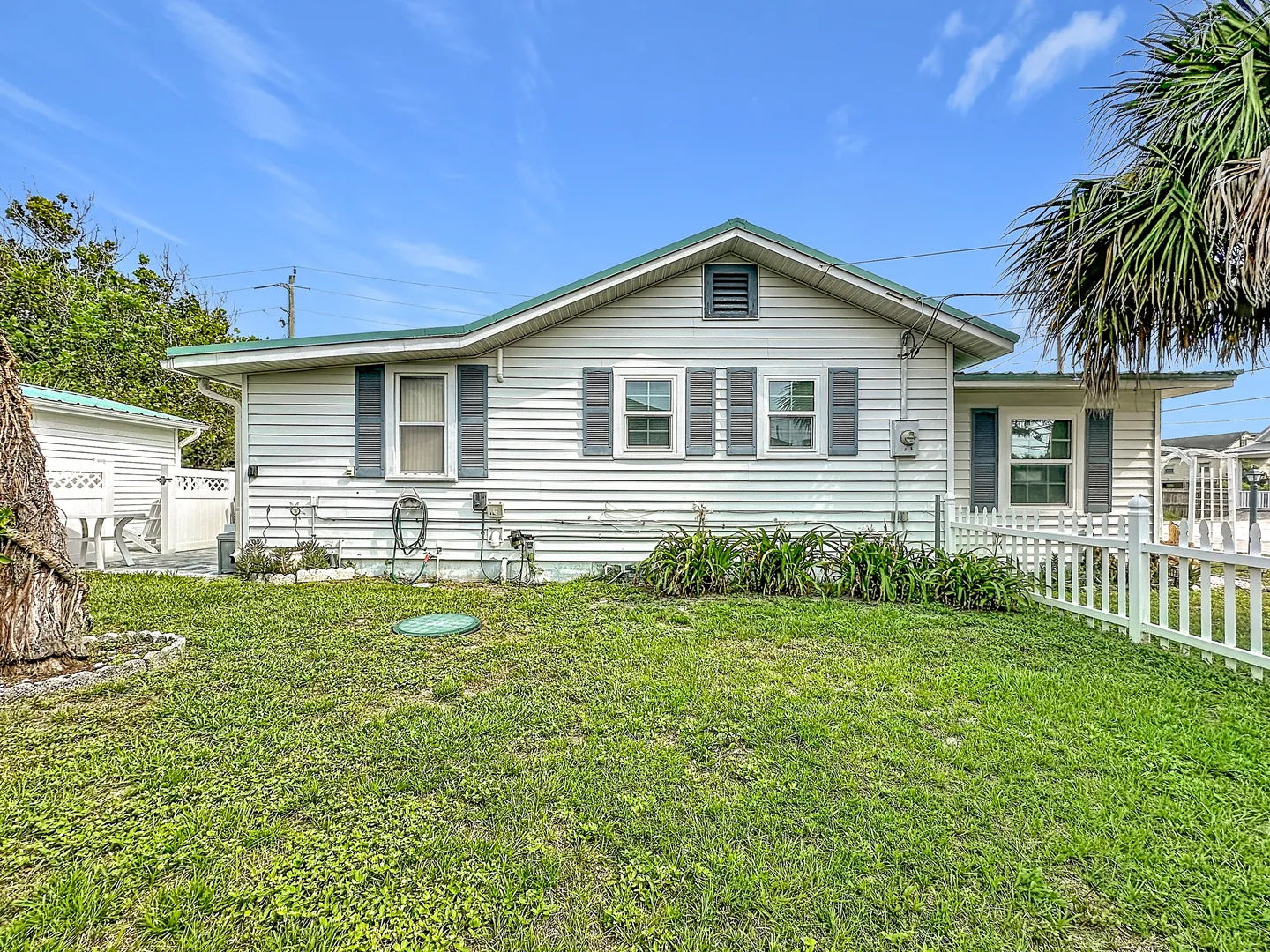 Exterior side view of a white single-story home with grey shutters, green trim, a grassy yard, and a white picket fence under a blue sky.