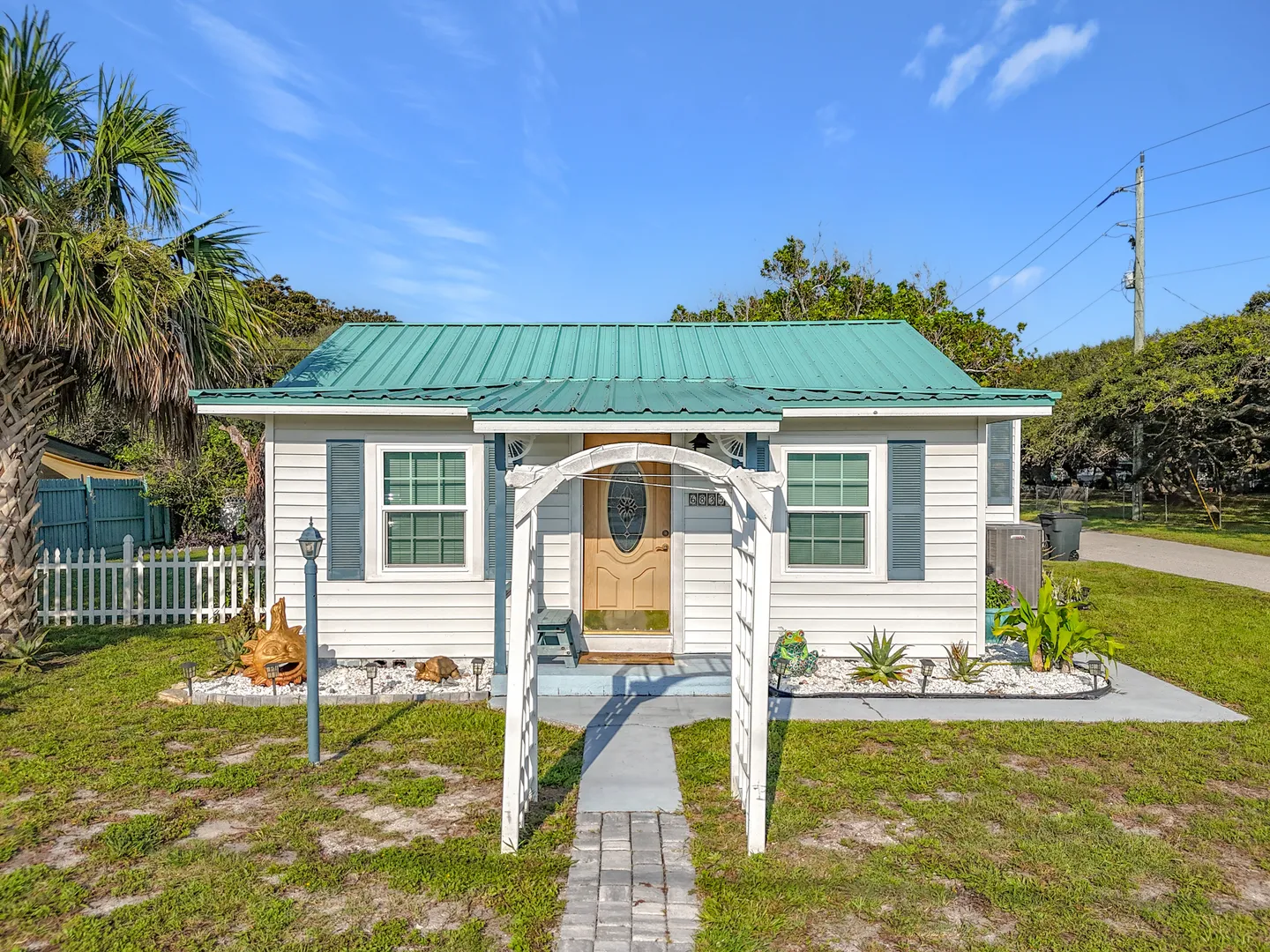 Front exterior of a small white cottage with a green metal roof, a white garden arbor, and landscaped front yard under a blue sky.