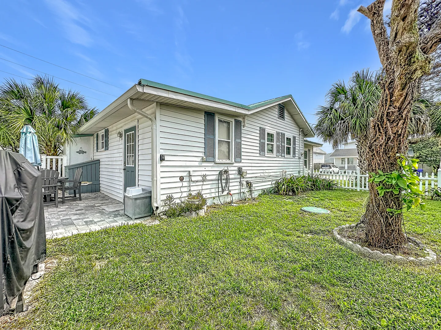 Exterior of a white single-story cottage with gray shutters, a grassy lawn, a tree, and a paved side patio.