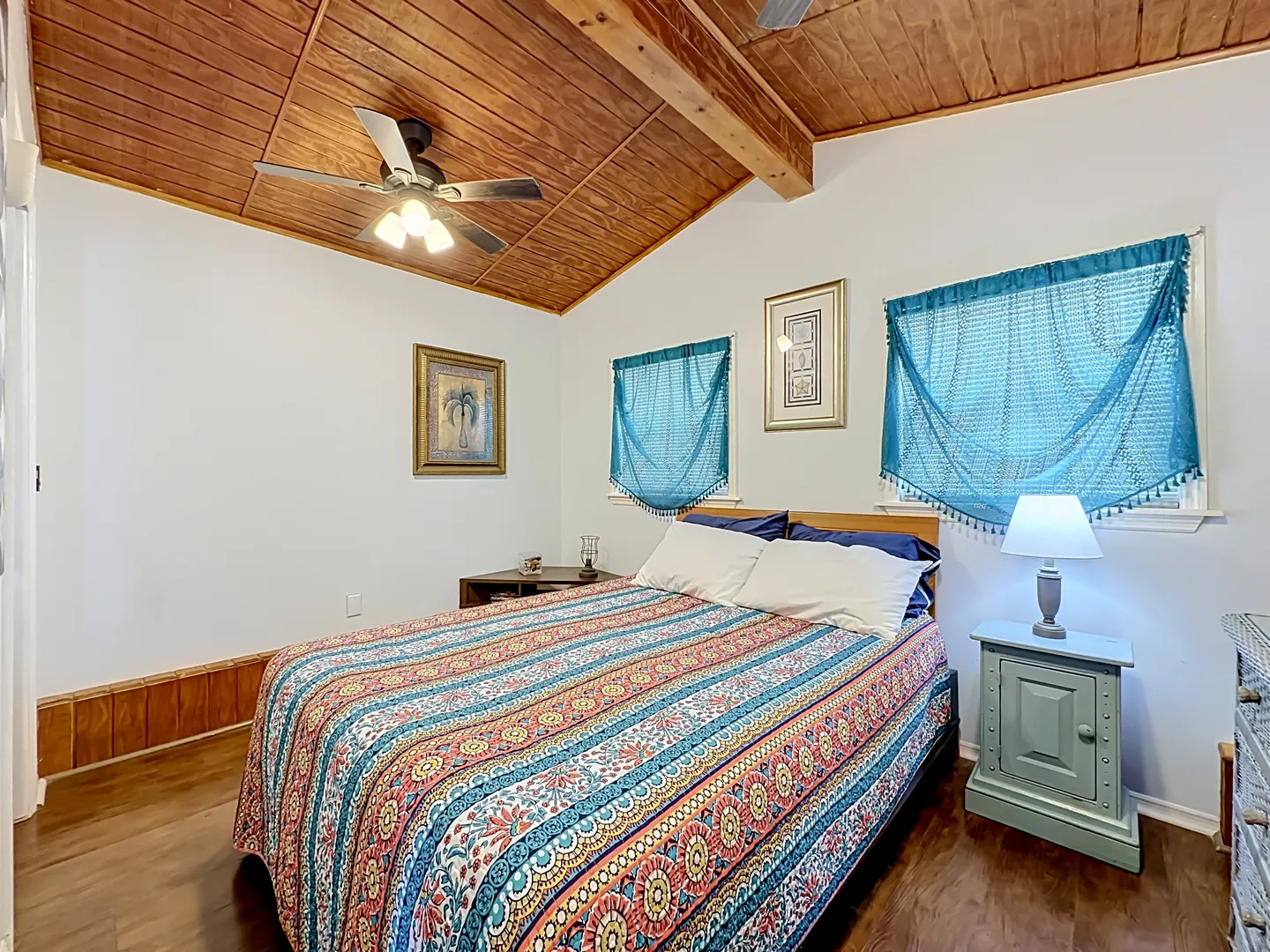 Bedroom with a patterned bedspread, wood plank ceiling with ceiling fan, and teal window coverings.