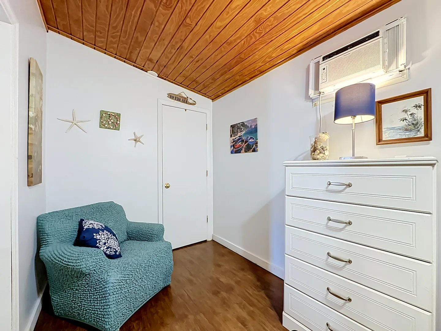 Corner of a bedroom featuring wood-paneled ceiling, teal armchair, white dresser, and coastal decorations.