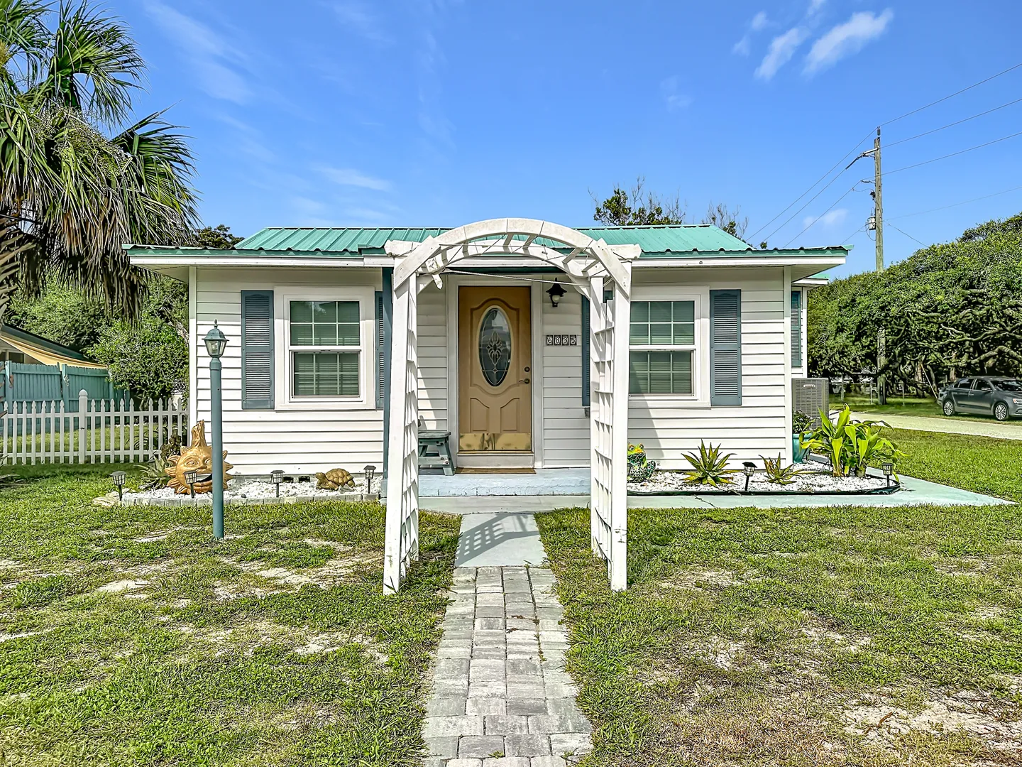 Exterior front facade of a small single-story white cottage with a teal metal roof, front yard arbor, and stone pathway.