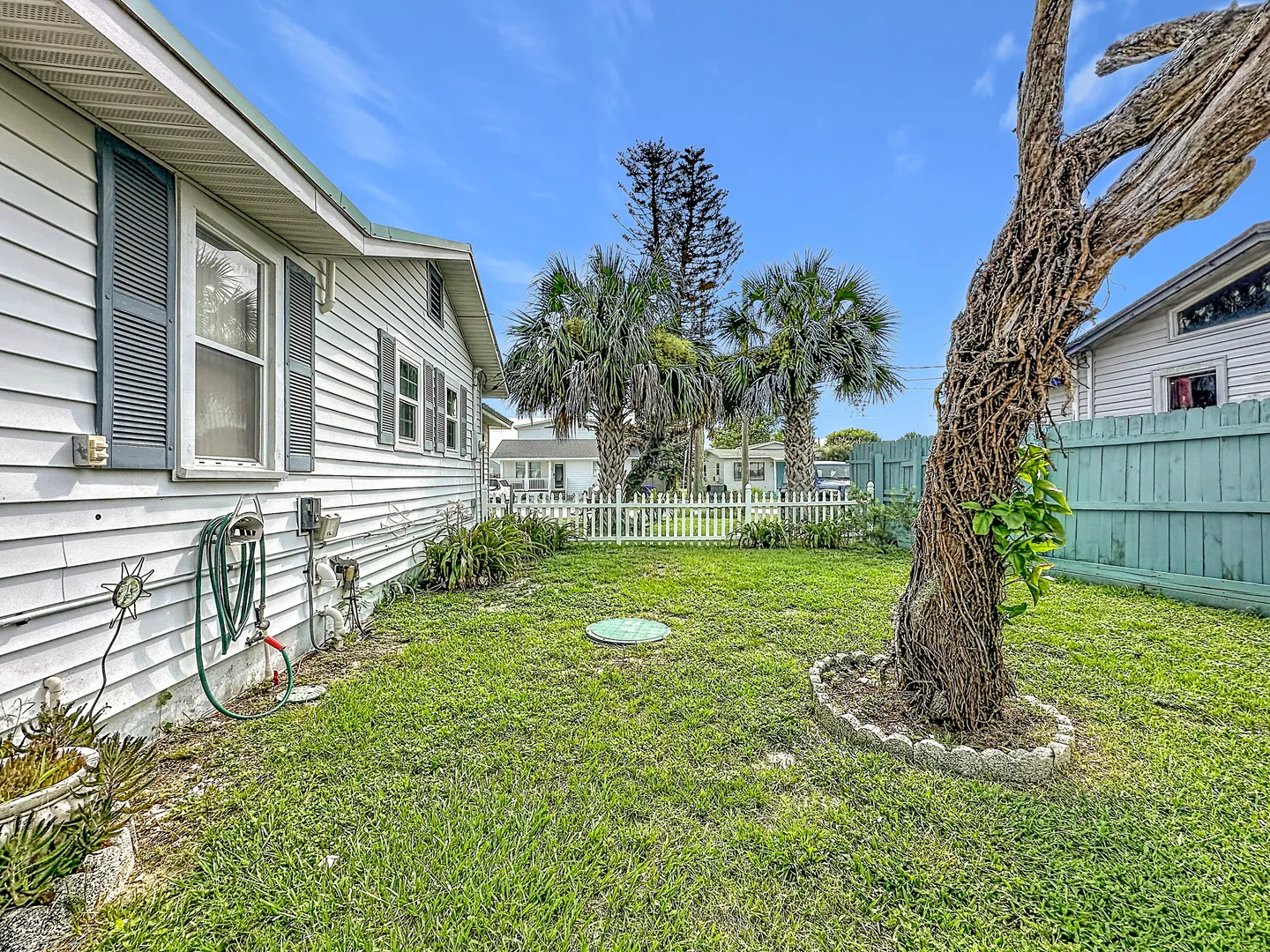 A grassy yard alongside a white home with siding, bordered by wooden and white picket fences with palm trees in the background.