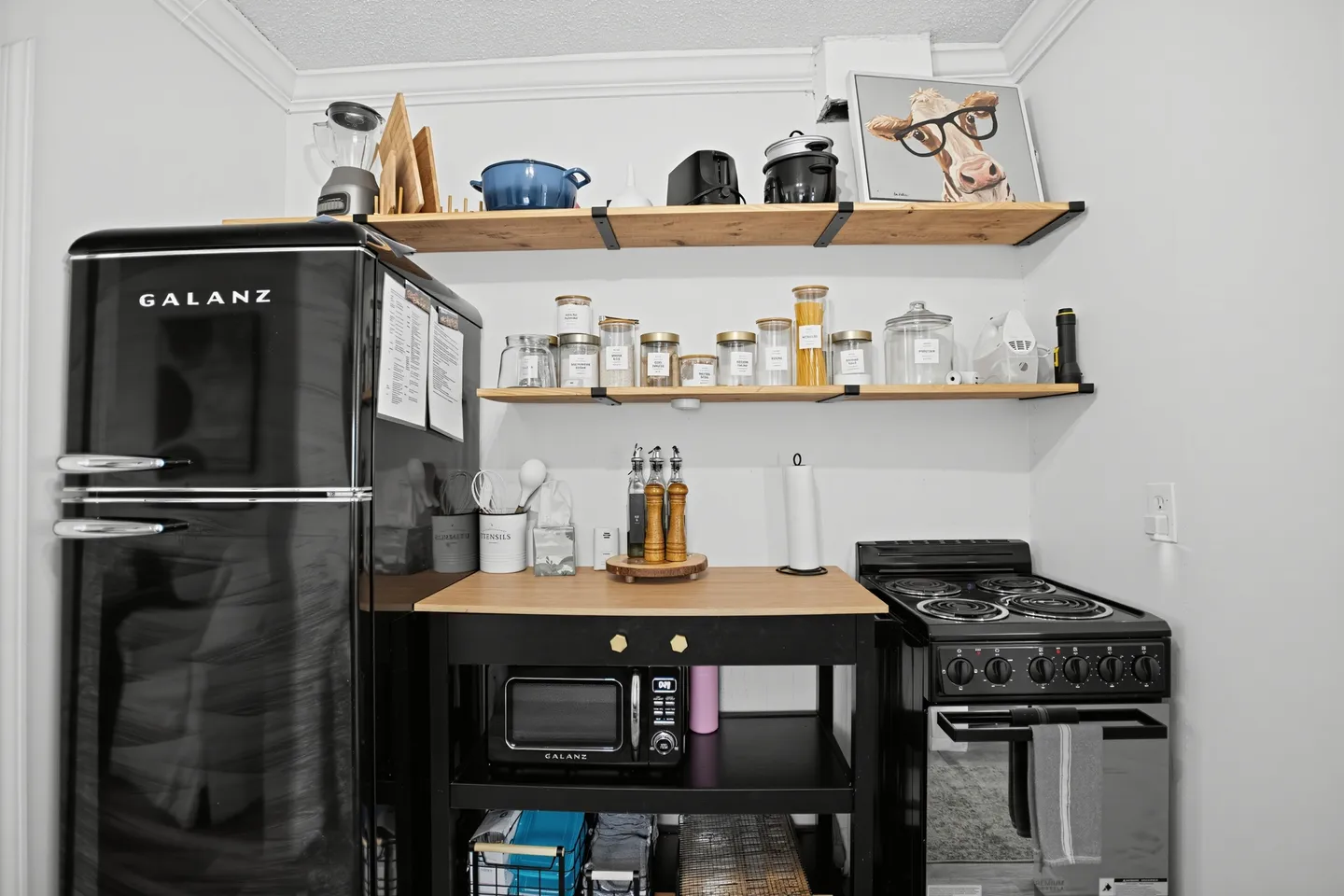 A compact kitchenette setup with a retro black refrigerator, microwave cart, small stove, and two floating wooden shelves.