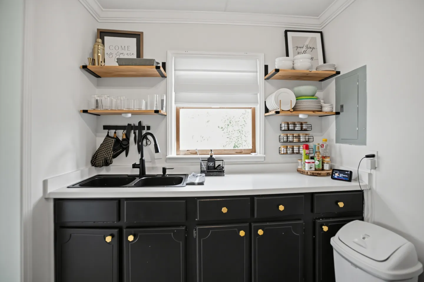 Kitchen counter area with black lower cabinets, white countertops, a double-basin black sink, and open wooden floating shelves displaying dishware.