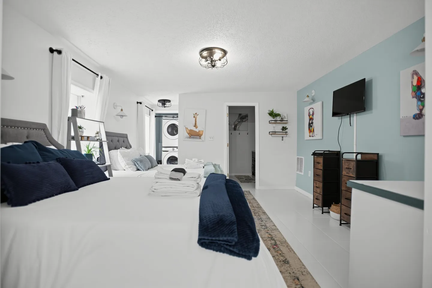 Bedroom with two beds, white linens, a light blue accent wall, a wall-mounted TV, and an open doorway showing a washer and dryer.
