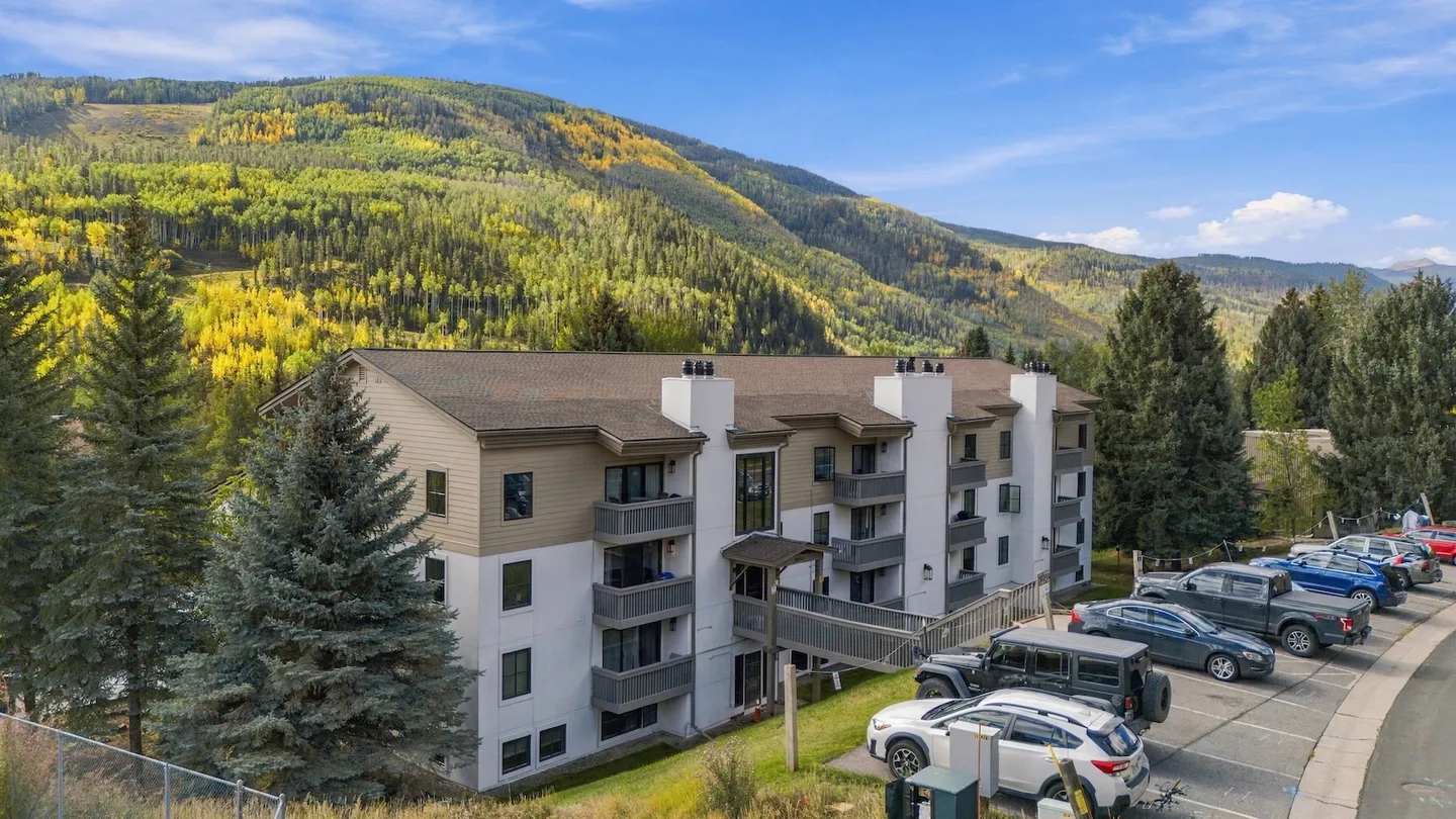 Exterior view of a multi-story condominium building backed by a mountain with fall foliage and parked cars in the foreground.