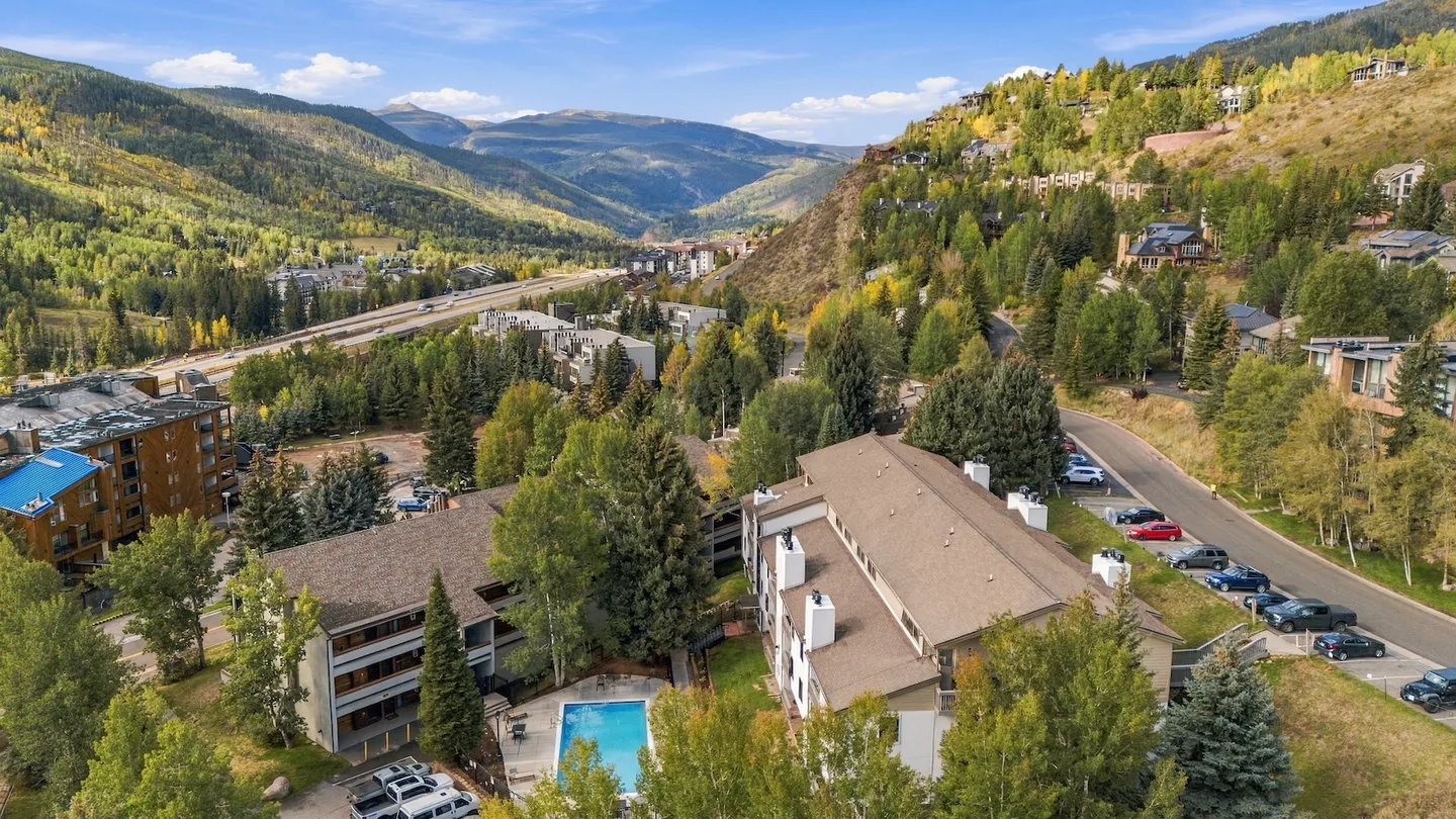 Aerial view of a mountain valley residential complex with an outdoor swimming pool, paved roads, and forested hillsides.