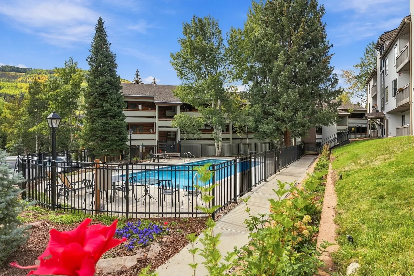 Outdoor swimming pool enclosed by a black metal fence, surrounded by multi-story residential buildings, trees, and landscaped gardens.