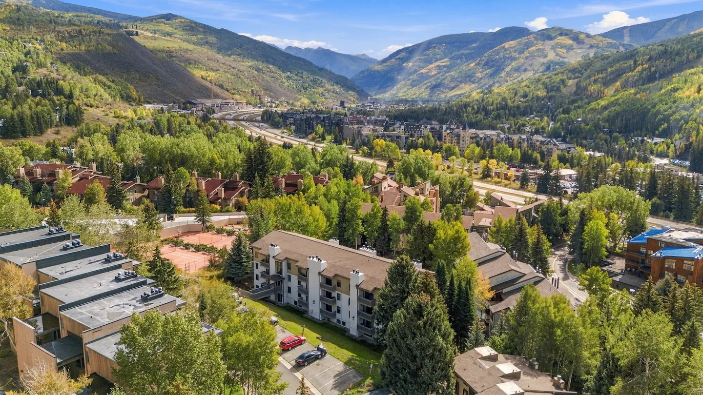 Elevated view of a mountain valley community with multi-story residential buildings, tennis courts, and forested mountains in the background.
