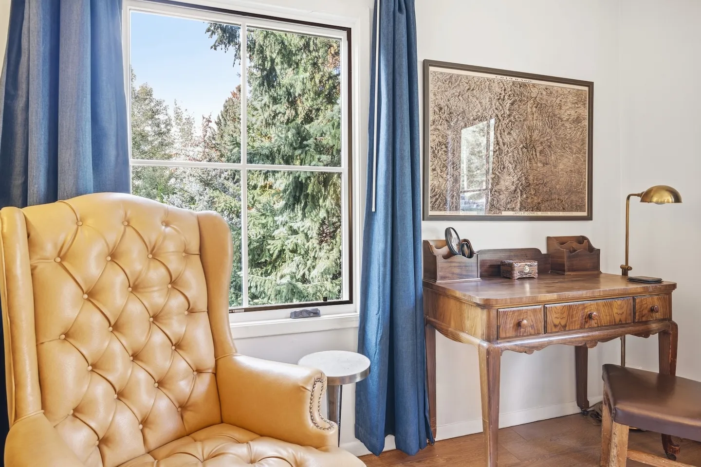 A leather tufted armchair next to a window with blue curtains and a vintage wooden desk with framed artwork above it.