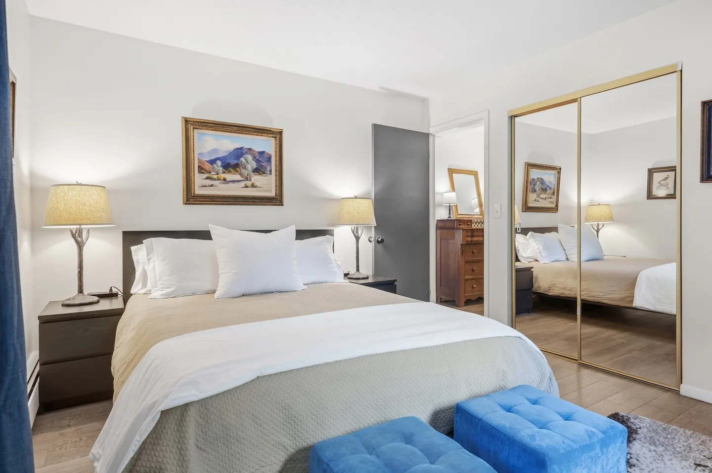 Bedroom with a made bed flanked by nightstands and lamps, mirrored closet doors, and two blue ottomans at the foot of the bed.