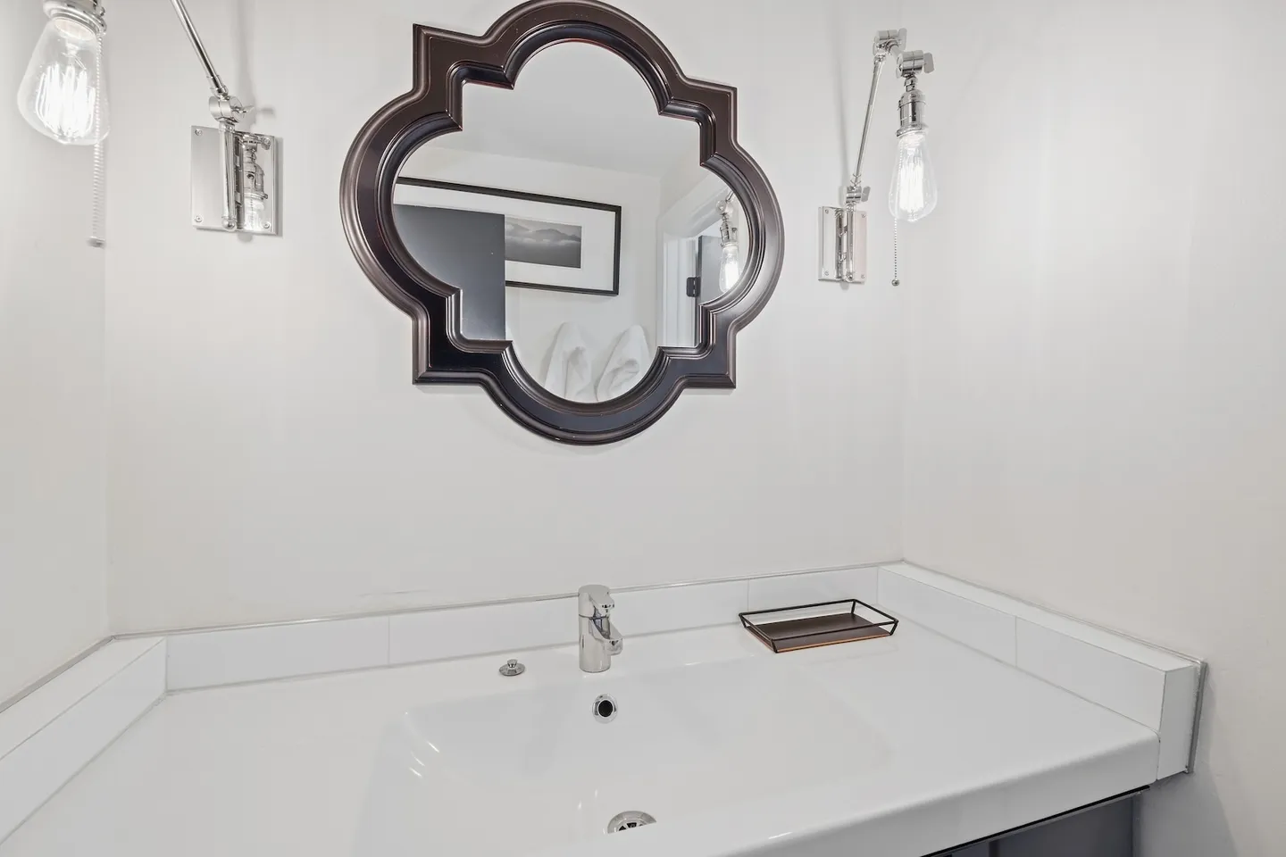 Bathroom vanity with a white countertop and sink, topped by an ornate dark-framed mirror and wall sconces.