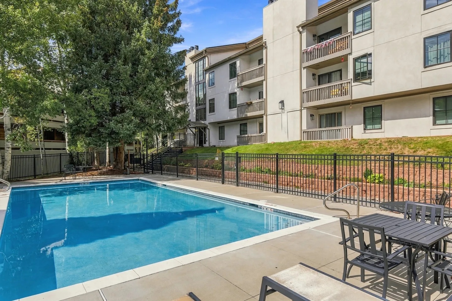 Outdoor rectangular swimming pool surrounded by a concrete patio and a black metal fence, with a multi-story apartment building and trees behind it.