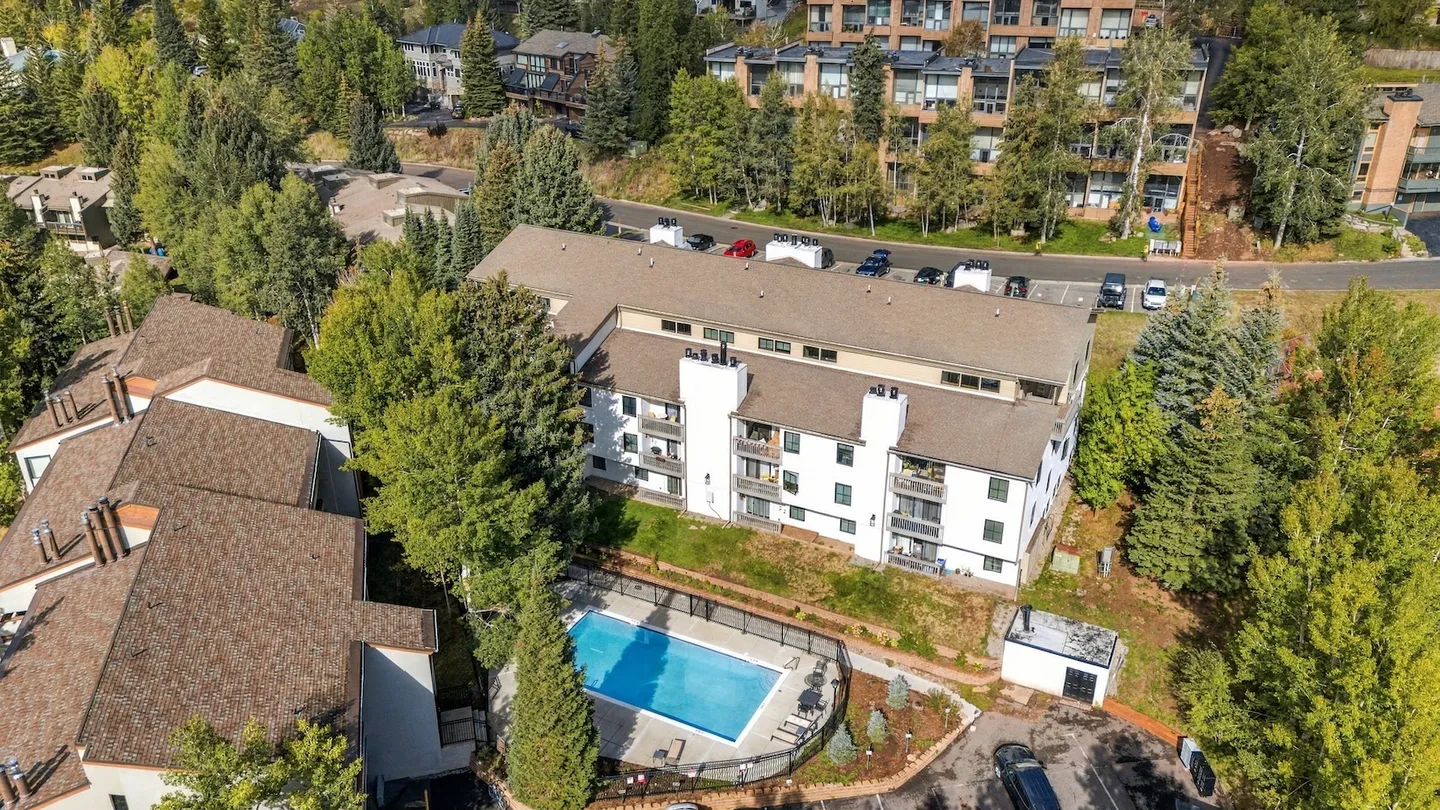 Aerial view of a multistory residential building featuring an outdoor swimming pool, parking areas, and surrounding green trees.