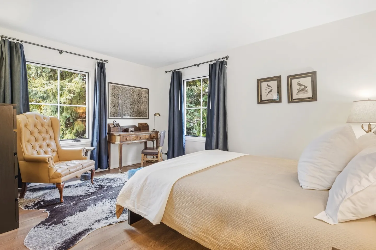 Bedroom featuring a bed with neutral linens, a tufted leather armchair, a small writing desk, and windows with dark curtains overlooking greenery.