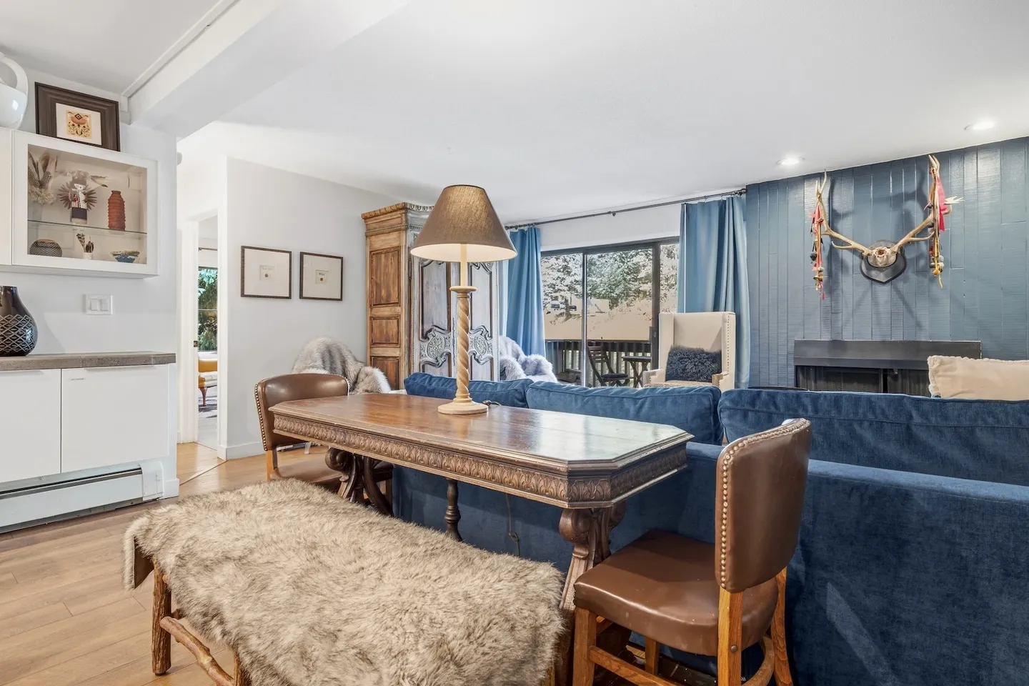 Living room featuring a long wooden console table behind a blue sofa, adorned with a bench, chairs, a lamp, and a blue accent wall with a mounted rack.
