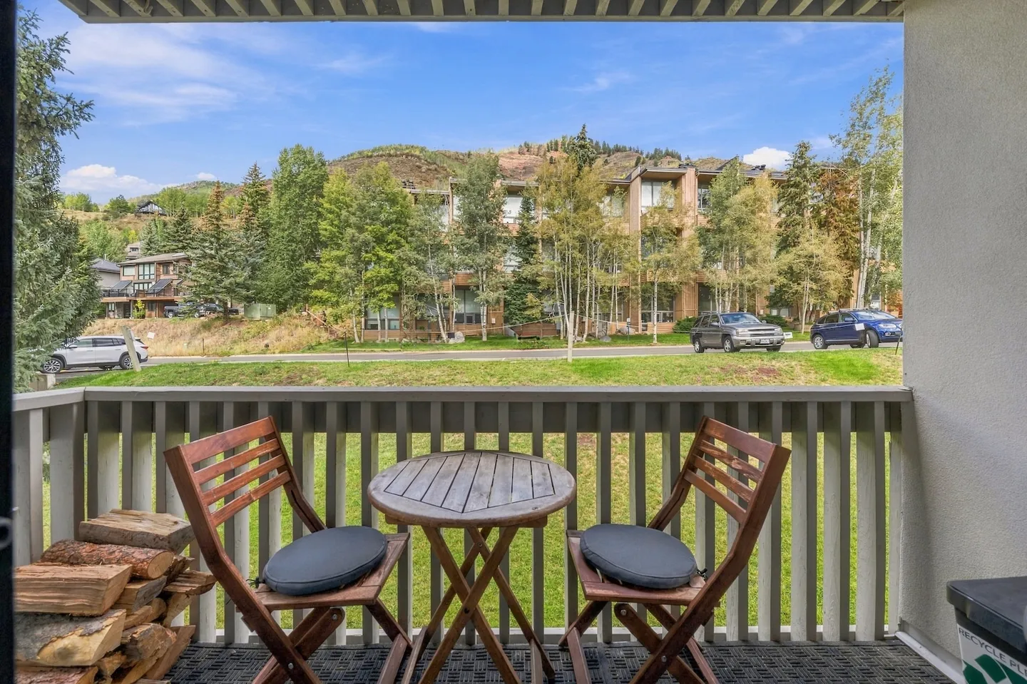 Balcony with two wooden folding chairs and a small round table overlooking a street and trees.