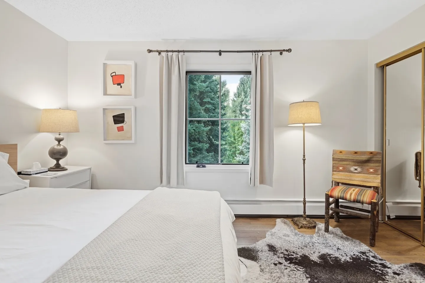 Bedroom with a bed in the foreground, a window looking out onto pine trees, a floor lamp, and a rustic wooden chair on a cowhide-style rug.