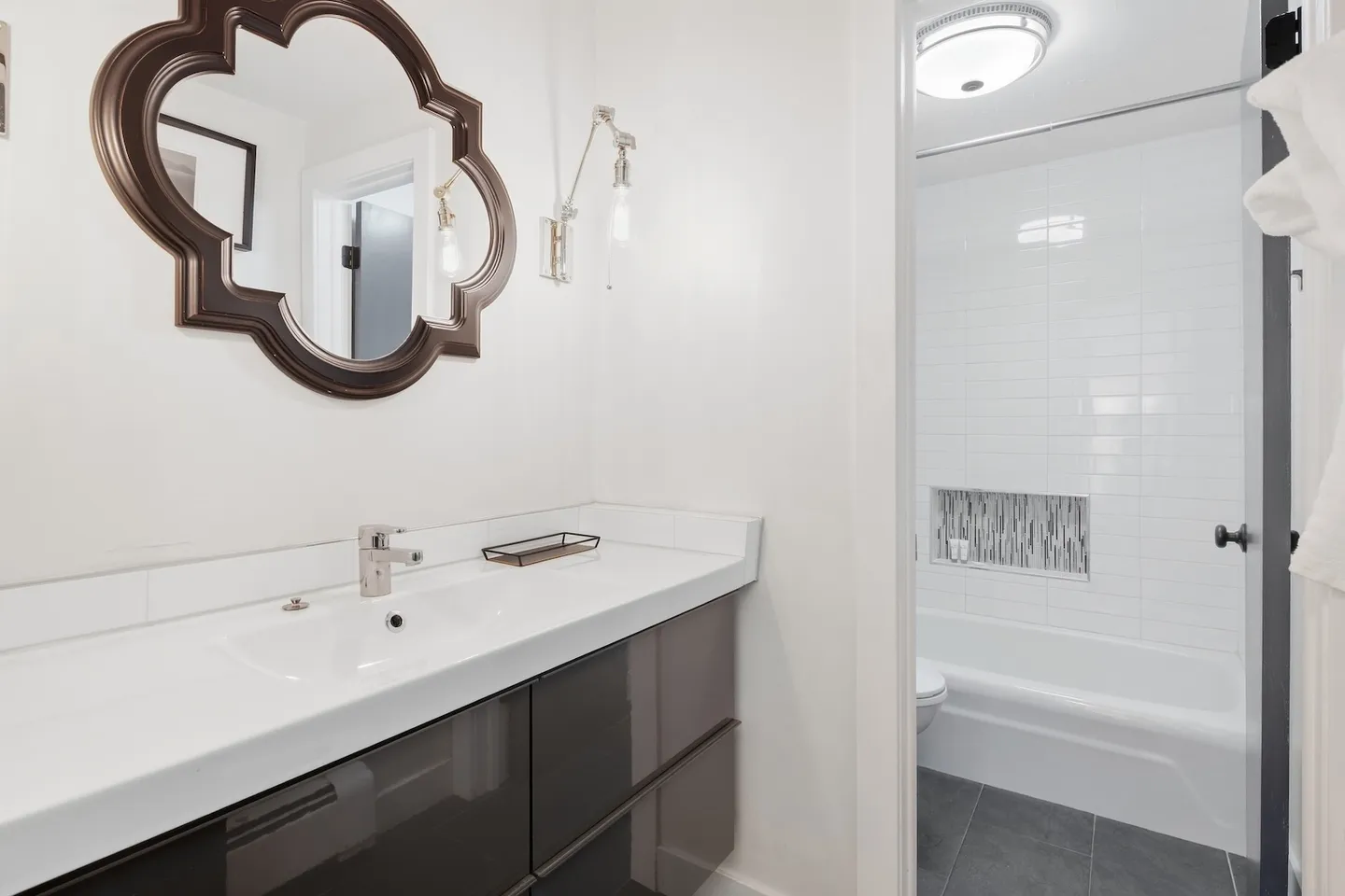 Bathroom with white countertop, dark glossy vanity drawers, decorative mirror, and a view into the tub-shower area.