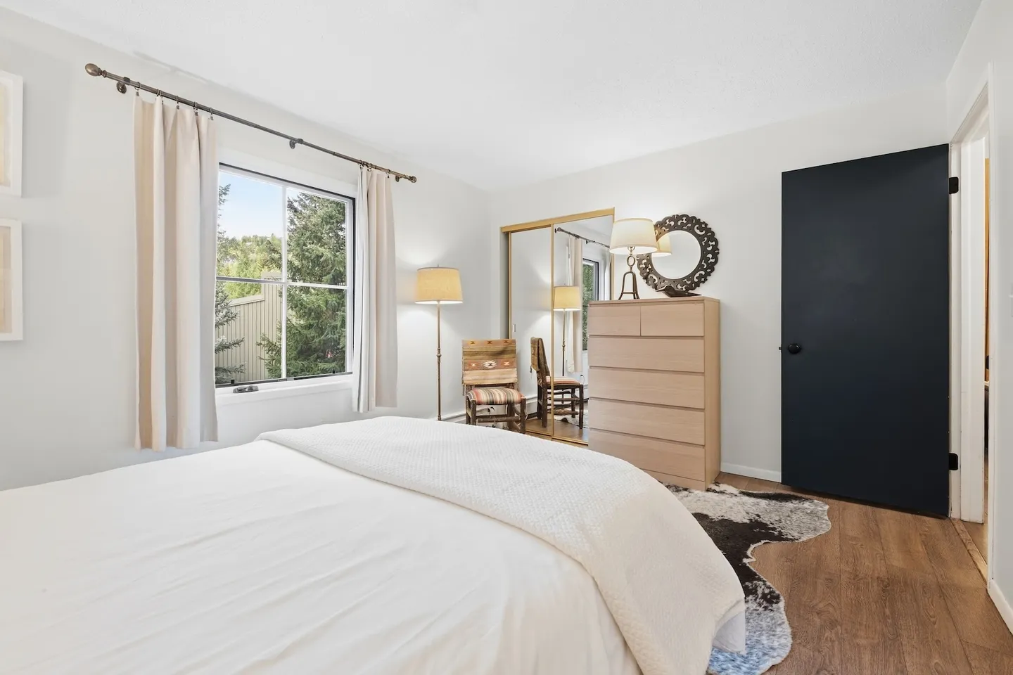 Bedroom with a bed made with white linens, a window with curtains, a wood dresser, and mirrored closet doors.