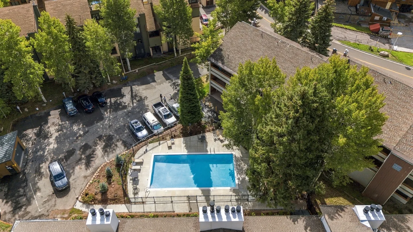 Aerial view of an outdoor community swimming pool surrounded by a fence, concrete deck, parking areas, and residential buildings.