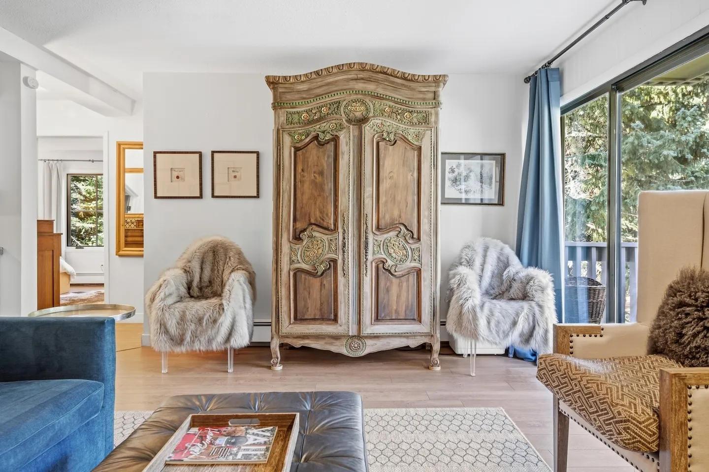A living room seating area featuring an ornate carved wooden armoire flanked by two faux-fur chairs next to sliding glass doors.