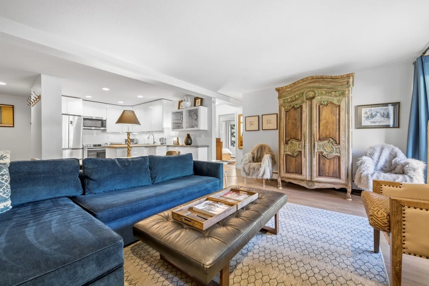 Open-concept living room with a blue sectional sofa, tufted leather ottoman, patterned rug, and antique wardrobe, leading into a kitchen with white cabinetry.
