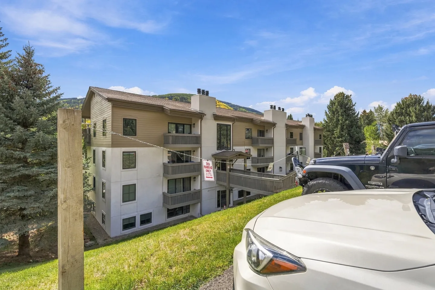 Exterior view of a multi-story condominium building with balconies, elevated walkway, and parked cars in the foreground.