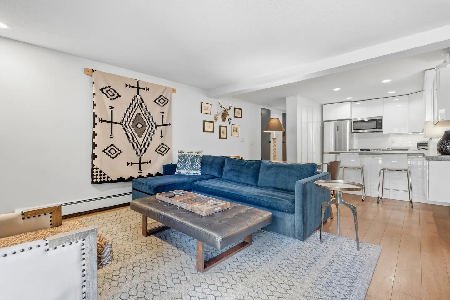 Open living room with a blue sectional sofa, patterned area rug, tufted ottoman, and view into the kitchen with white cabinetry.