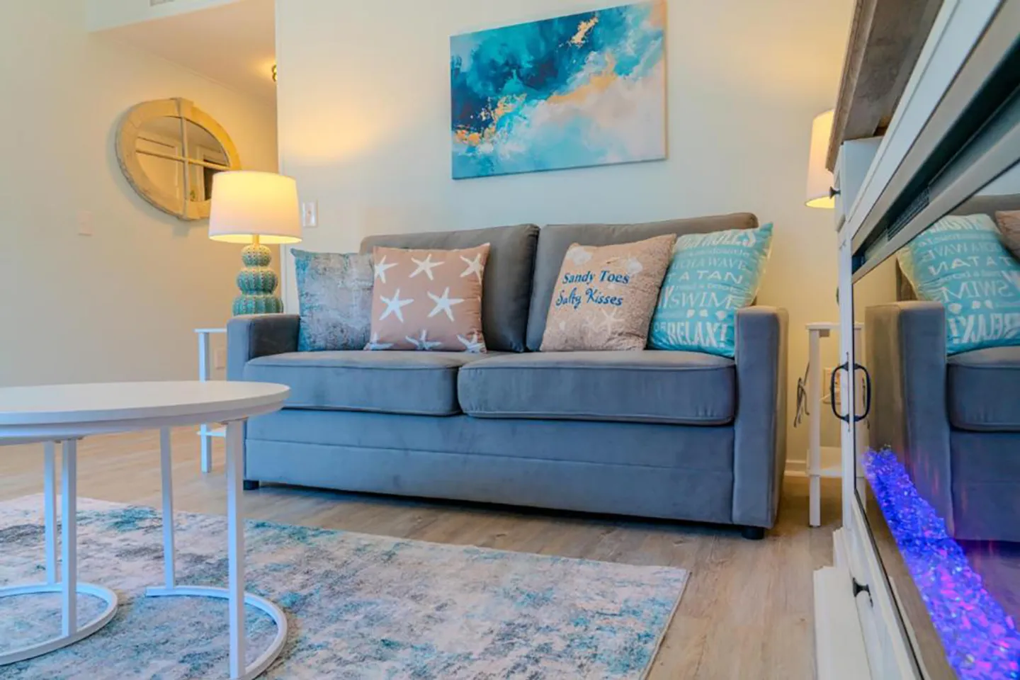 Living room with a grey couch, coastal decorative pillows, an electric fireplace with blue lights, and an abstract blue painting on the wall.